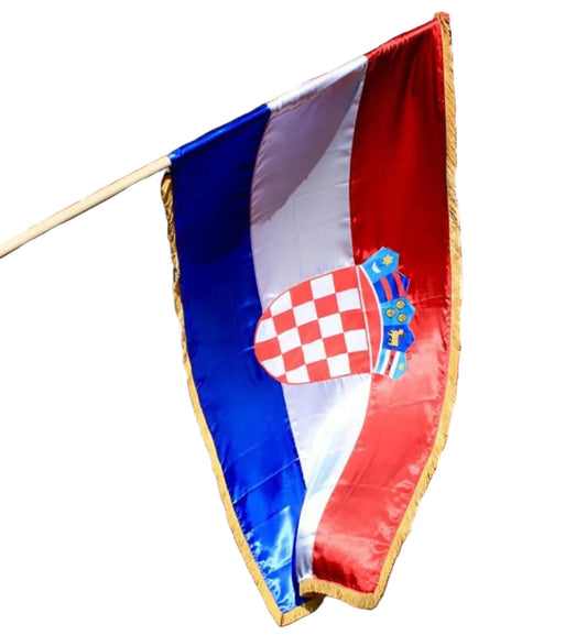 A Croatian flag hanging from a pole outdoors in front of a black metal fence and building entrance. The flag has the characteristic red, white, and blue vertical bands with the Croatian coat of arms near the hoist side and a gold fringe along the edge. Official Croatian Silk Flag 200cm x 100cm on snc shopncro