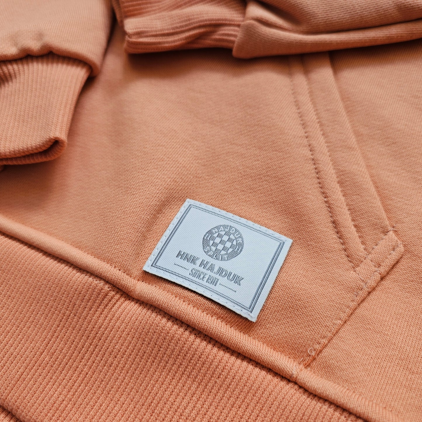 Close-up of a peach-colored hoodie with a brand label on the chest pocket. Kids Tracksuit Hajduk
