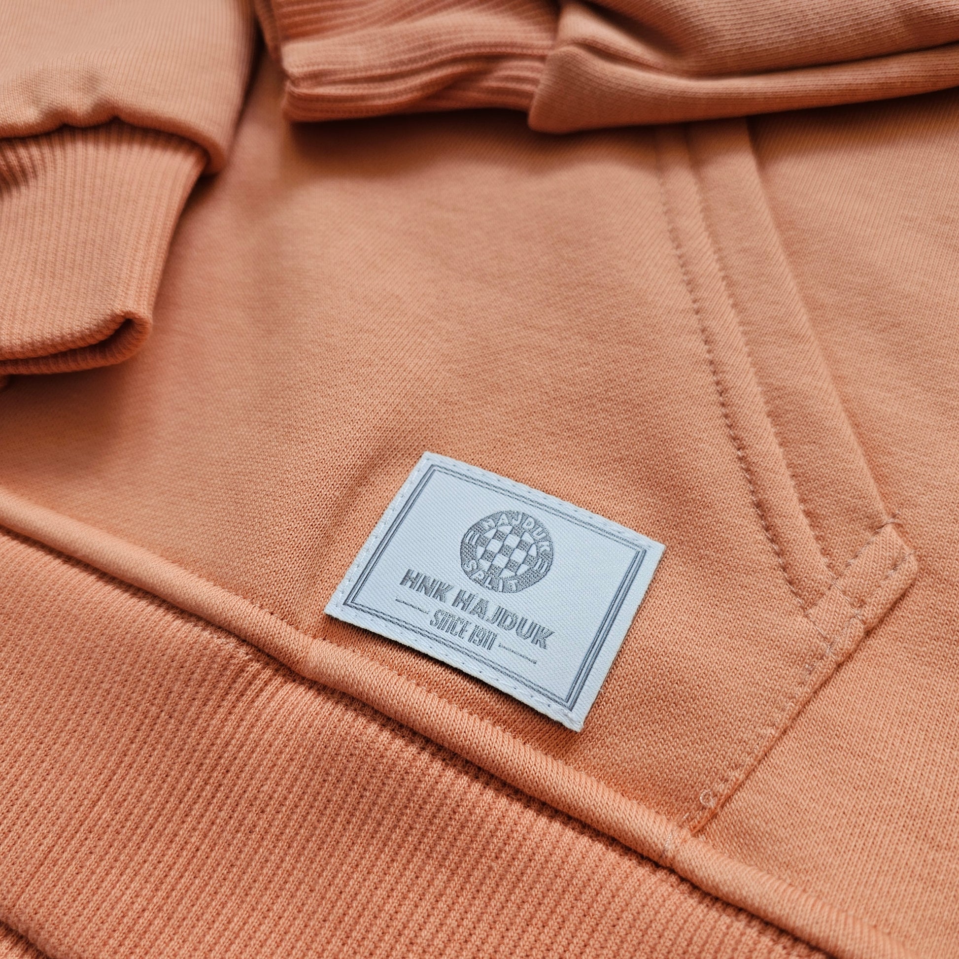 Close-up of a peach-colored hoodie with a brand label on the chest pocket. Kids Tracksuit Hajduk